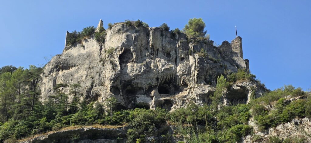 A tall pale limestone cliff above Fontaine-de-Vaucluse riddled with caves and hollows, with the broken walls and a small tower of the ruined bishops castle standing along the top and pines below