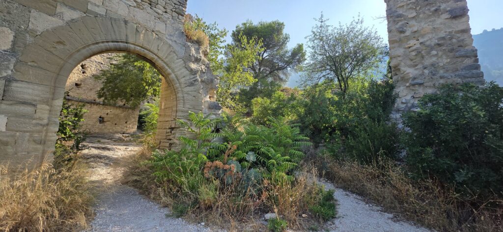 A surviving stone archway in a ruined wall above Fontaine-de-Vaucluse, gravel path running through it and dry grass, ferns and scrub growing over the fallen stonework