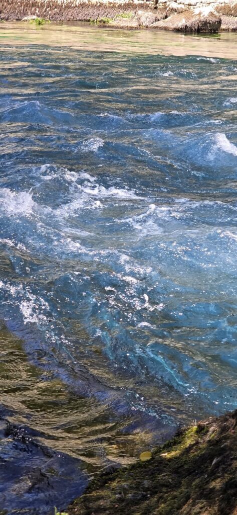 Fast turquoise water of the Sorgue breaking white over a shallow rocky ledge, with green weed and stones visible through the clear water at the edge
