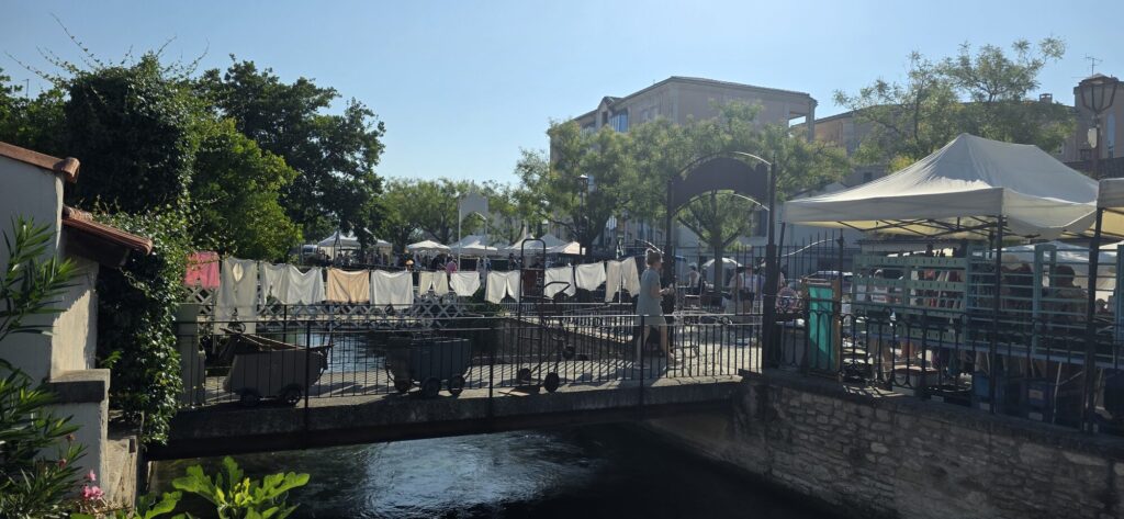 A branch of the Sorgue running under an iron footbridge at L'Isle-sur-la-Sorgue with old white linen sheets pegged along the canal railings and white market awnings behind
