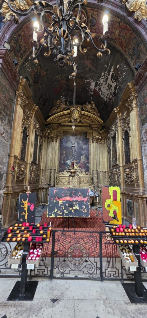 A side chapel in a baroque church with a heavily gilded carved retable and an old painting at its centre, a painted vault above, an iron chandelier, and three modern abstract paintings in red, black and yellow standing on easels in front of the altar with racks of votive candles either side