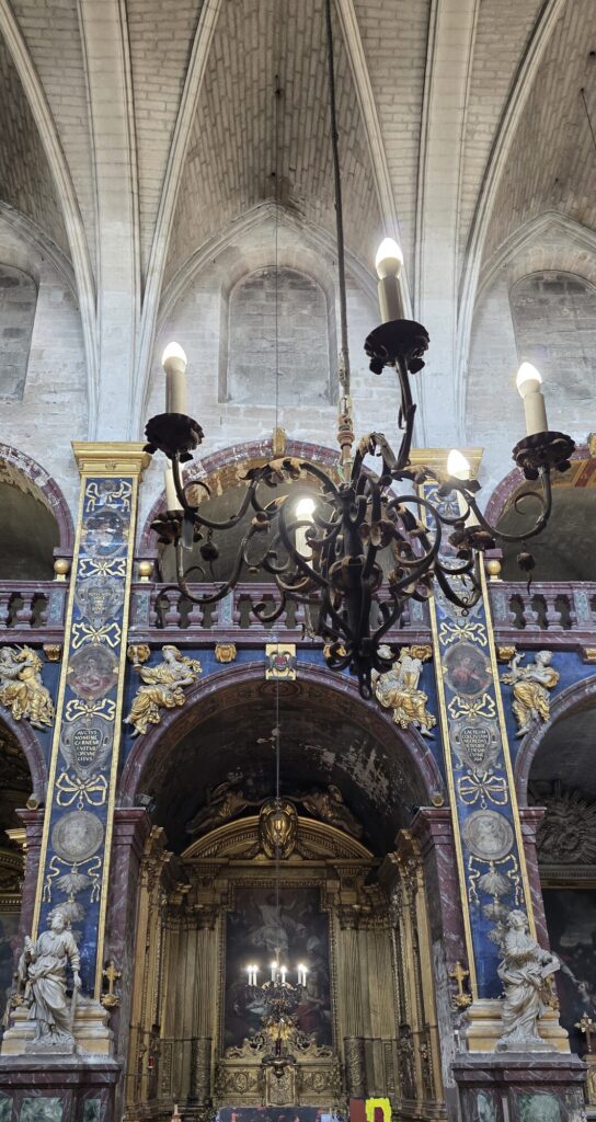Looking up in the church nave at bare pale Gothic stone vaulting above, and below it a band of dark blue and gold baroque decoration with gilded angels, painted medallions and white statues, with a black iron chandelier hanging in the middle