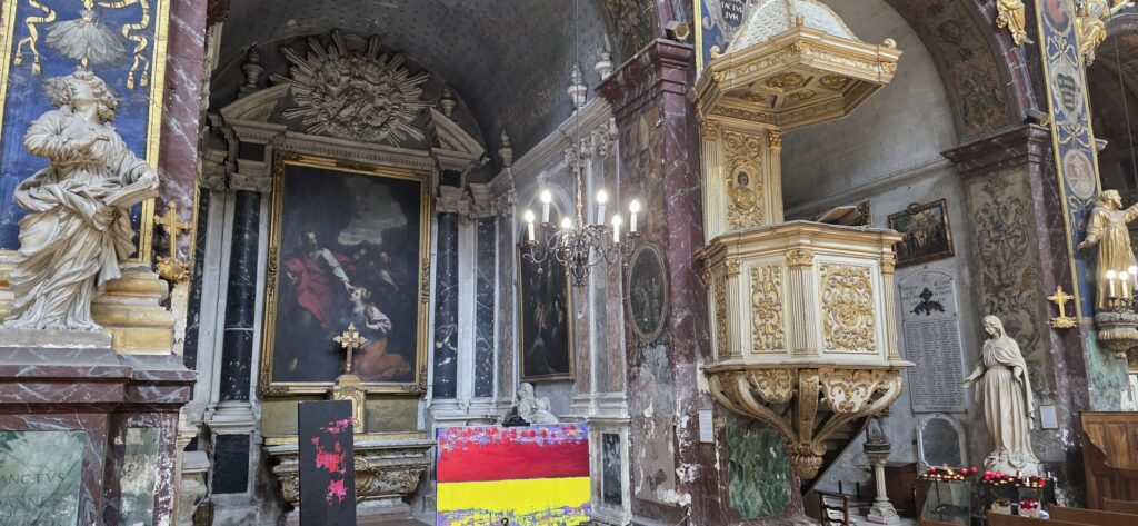 Wide view inside the baroque church showing a carved gilded pulpit, marble pilasters, white stone statues in niches, a large old master canvas in a gold frame, and a big red and yellow abstract painting standing on the floor beside the altar step