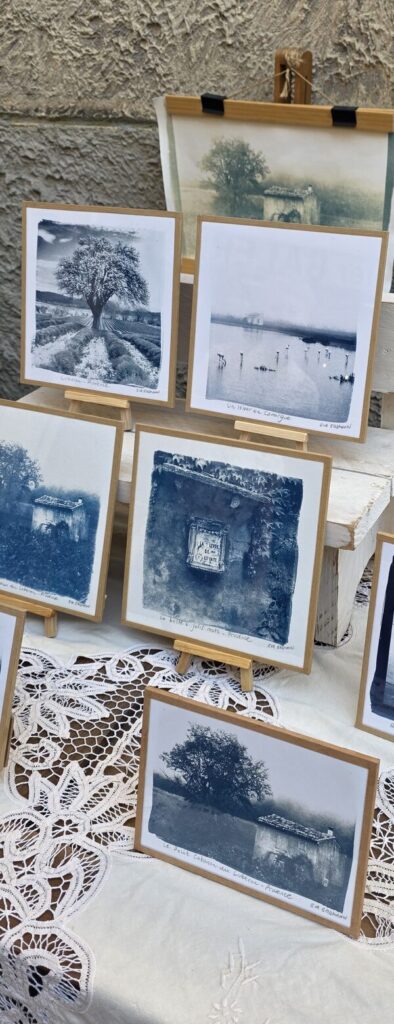 Five blue cyanotype prints of Provence landscapes propped on small wooden easels on a lace tablecloth in the street