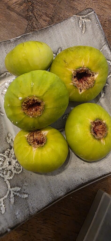 Four green figs with split brown eyes sitting on a patterned white plate on a dark wooden table