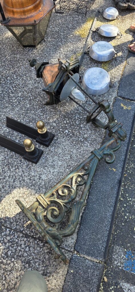 A large green patinated wrought iron lantern bracket with scrolled curves lying on the pavement beside copper pans, an old lantern and iron gate finials