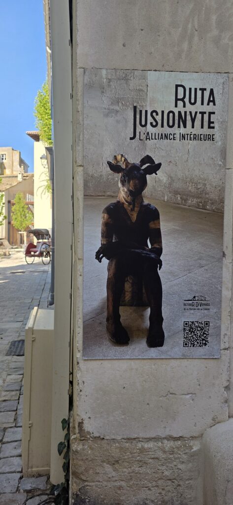 Exhibition poster on a stone corner in L'Isle-sur-la-Sorgue for Ruta Jusionyte, L'Alliance Interieure, showing a dark seated sculpture with a stag's head and human hands