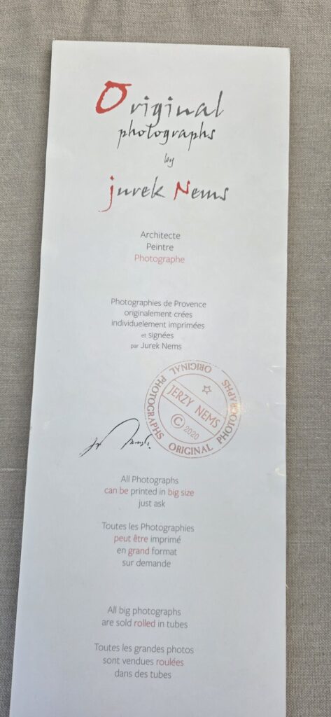 A tall printed leaflet reading Original photographs by Jurek Nems, architecte peintre photographe, with a red circular authenticity stamp and a signature, laid on grey linen