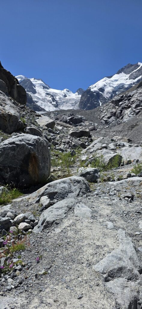 Looking up a wide valley of bare grey rock and boulders towards a glacier tongue and snow covered peaks under a deep blue sky, with no vegetation anywhere on the valley floor