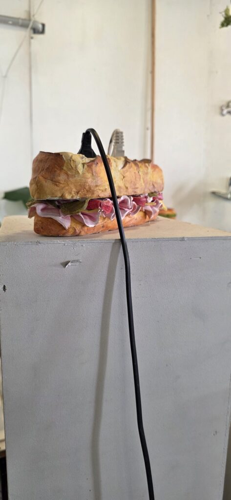 Sculpture of a filled baguette with ham, pickles and salad, modelled and painted, sitting alone on a white plinth with a black electrical cable running past it