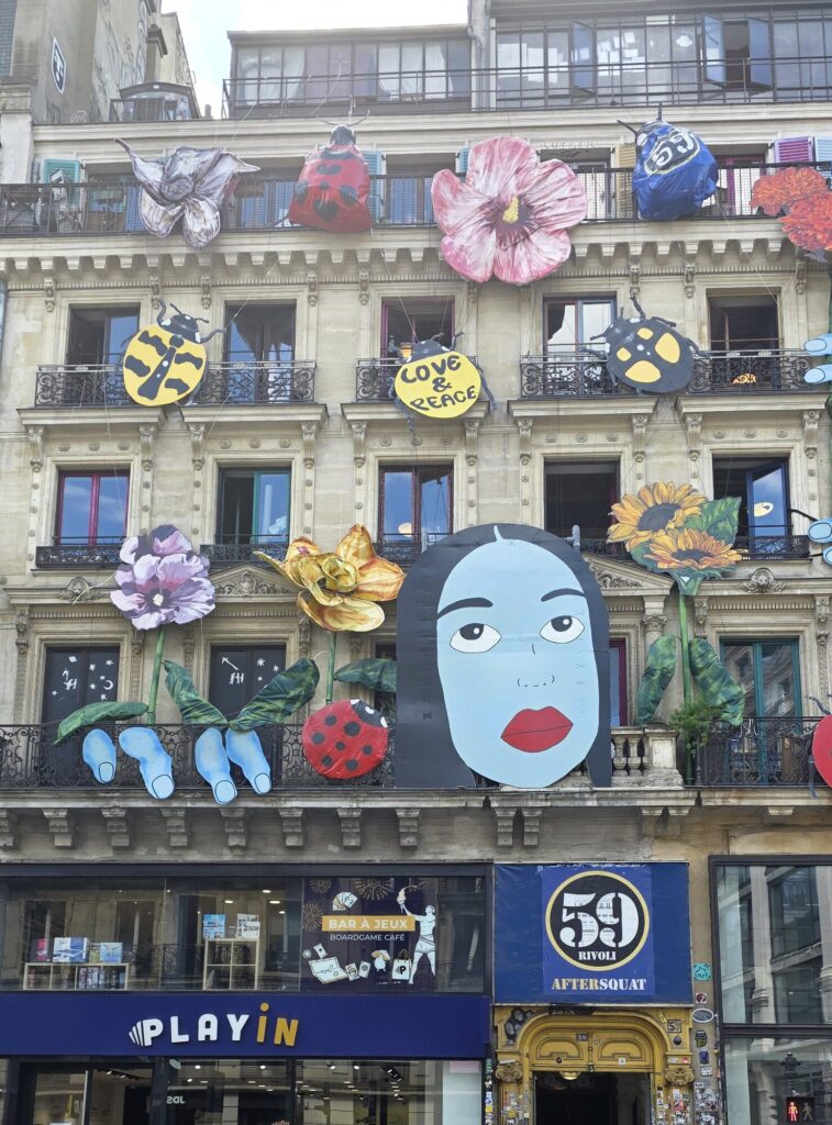 Classic Paris stone facade covered in enormous painted cut-out flowers, ladybirds, beetles and a giant flat face with red lips hung from the balconies, with a blue sign reading 59 Rivoli Aftersquat above the sticker-covered doorway