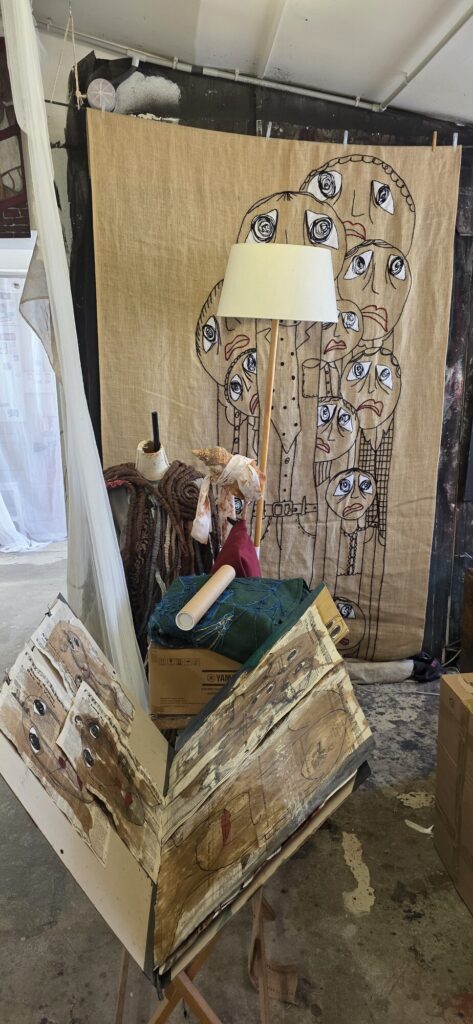 Studio with a large hessian sheet pegged up, covered in a crowd of outlined faces with huge staring eyes, and in front of it an enormous open book made of stitched printed pages carrying more drawn faces, propped on a wooden stand