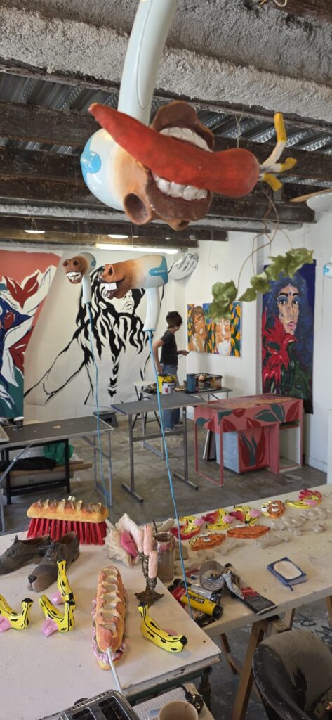 Bright studio with painted hairdryers hung from the ceiling on cords, each one fitted with a sculpted mouth full of teeth, a long table of painted bread and banana sculptures below, and a black ink figure painted straight onto the far wall