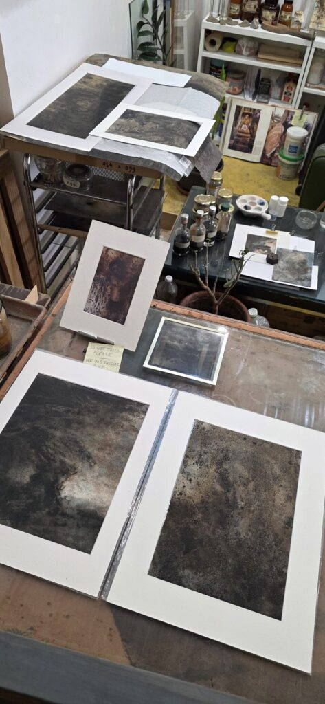 Printmaking studio, mounted dark tonal prints laid out on the steel bed of a press with tissue interleaves, small bottles of ink behind, a handwritten note reading do not touch please ne pas toucher svp