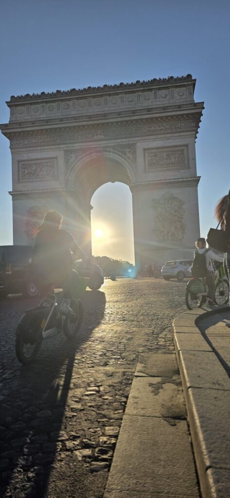 Riders on bicycles seen from behind heading up the avenue towards the Arc de Triomphe with the sun flaring through the arch ahead of them