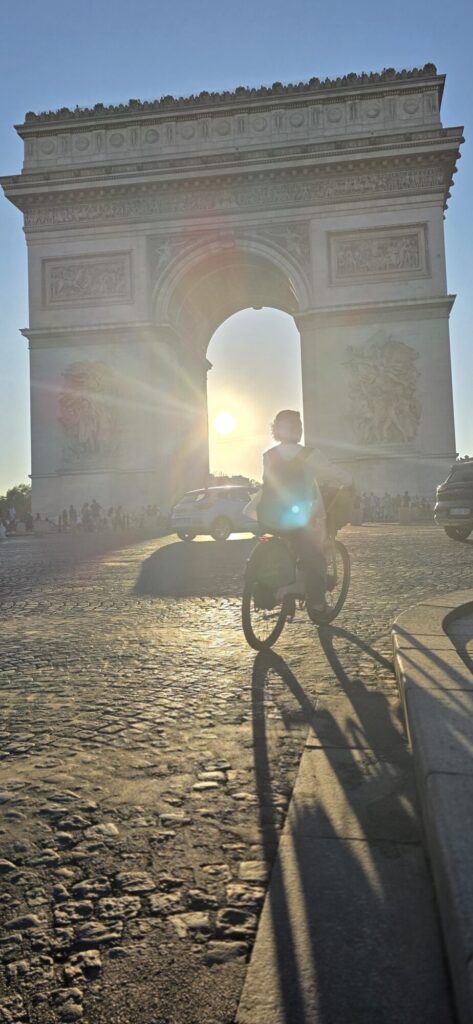 The Arc de Triomphe at golden hour with the low sun blazing directly through the arch and a single backlit cyclist crossing in silhouette