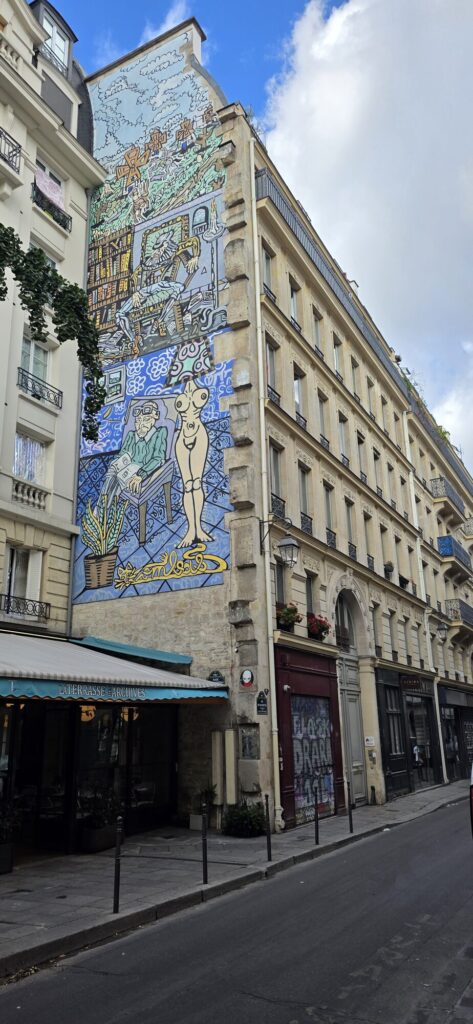 Tall Paris apartment building with a huge painted mural covering the whole side wall, showing a seated figure at a drawing board in heavy black outline and flat blue and ochre colour, with a cafe awning below