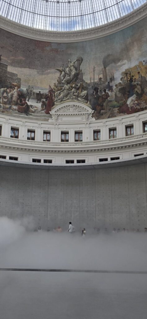 Interior of the Bourse de Commerce rotunda, a huge nineteenth century painted frieze curving under a glass dome, and below it a wall of white fog lying across the floor with distant visitors standing waist deep in it