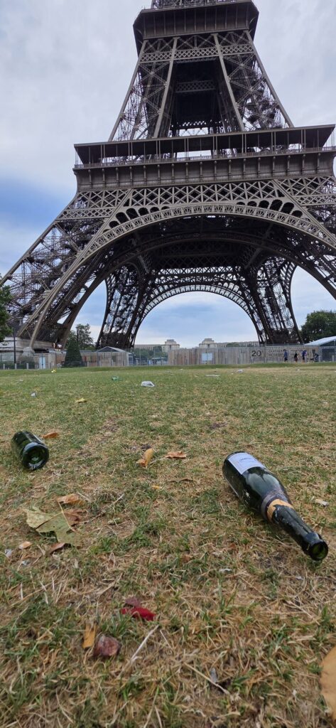 Two empty green champagne bottles lying on dry grass on the Champ de Mars with litter scattered around them and the base of the Eiffel Tower filling the sky behind