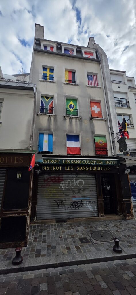 Narrow Paris building with eleven national flags hung in its windows above a bistro awning, roller shutter covered in tags, cobbled street below