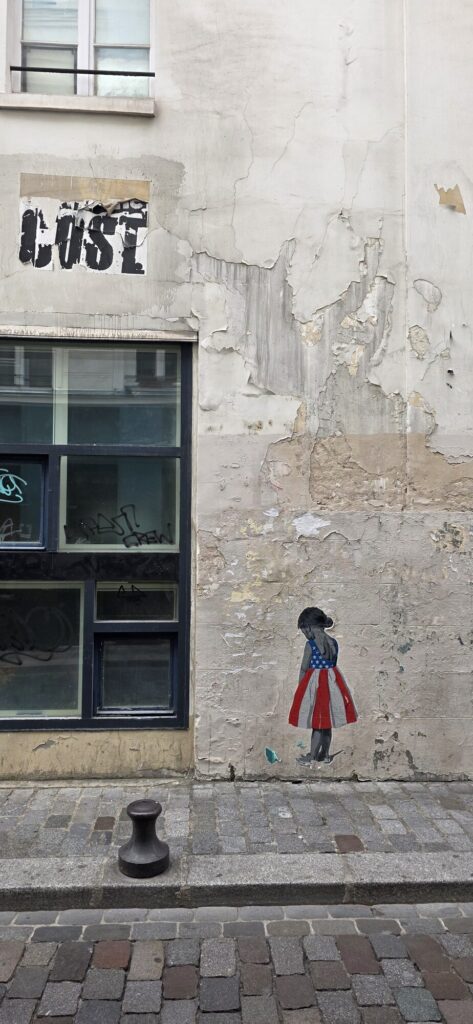 Paste-up of a small girl in a dress made from the stars and stripes, standing facing a peeling render wall beside a barred window, cobbles in the foreground