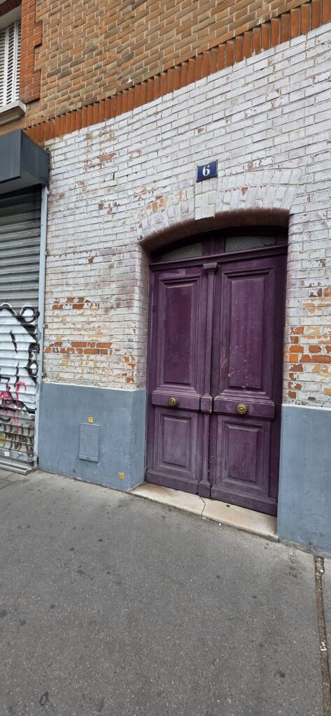 Tall aubergine painted double door with brass handles set in whitewashed brick under a red brick course, a blue enamel number six above it, tagged roller shutter to the left