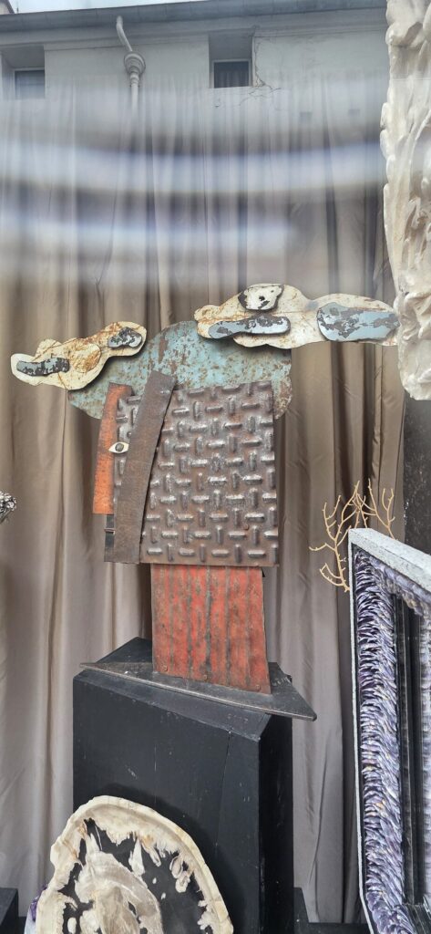 Gallery window seen through glass, holding a standing figure built from rusted steel plate and painted metal offcuts with cloud shapes for a head, beside a fossil slice and a purple mineral frame