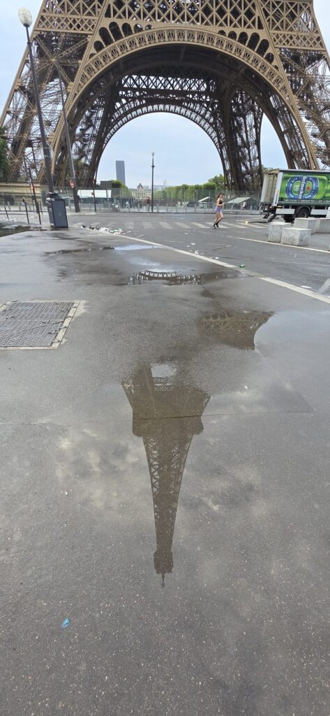 A wide empty road under the arch of the Eiffel Tower with the whole tower reflected upside down in a long shallow puddle, litter scattered across the tarmac and a delivery truck parked beyond