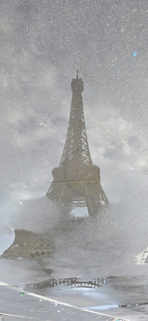 The Eiffel Tower reflected upside down in a shallow rain puddle on grey Paris paving, the reflection broken up by grit and cloud