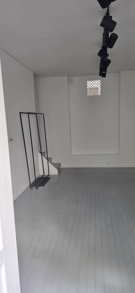 Small empty white gallery space with a track of black spotlights on the ceiling, grey painted floorboards, a short flight of steps at the back and a metal handrail leaning against the wall
