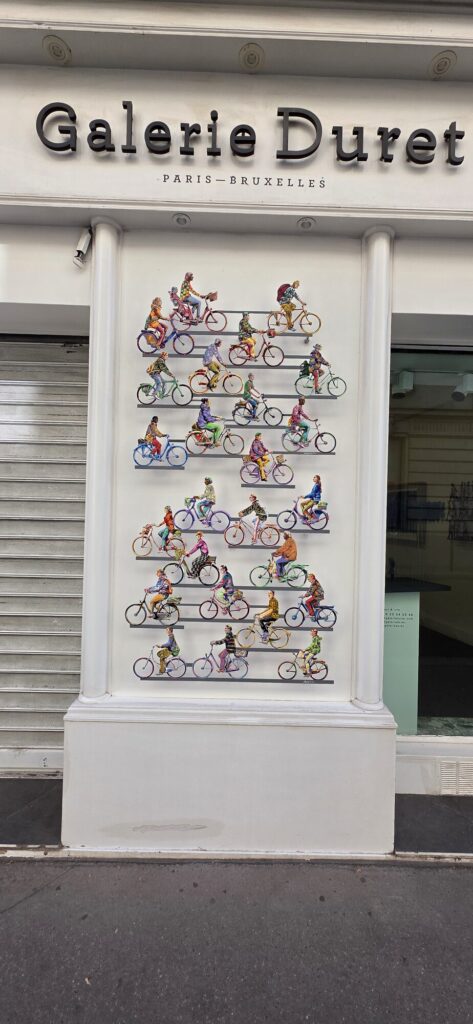 White storefront of Galerie Duret with Paris Bruxelles lettering and a wall sculpture of dozens of small painted cyclists mounted on the front