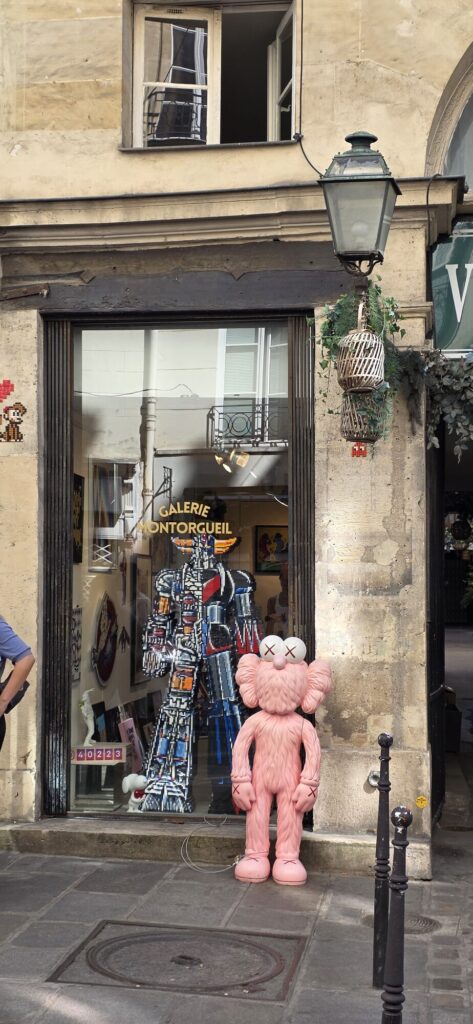 Galerie Montorgueil window with the pink furry KAWS figure beside a large pixel mosaic samurai style figure in blue and orange tiles, small artworks visible inside