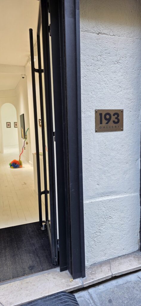 Open street door of a gallery with a brass plate reading 193 Gallery, looking through into a white space with pale floorboards, framed works on the walls and a brightly coloured beaded sculpture on the floor