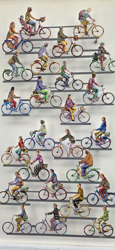Close-up of a David Gerstein painted steel wall sculpture showing rows of colourful cyclists riding across horizontal bars