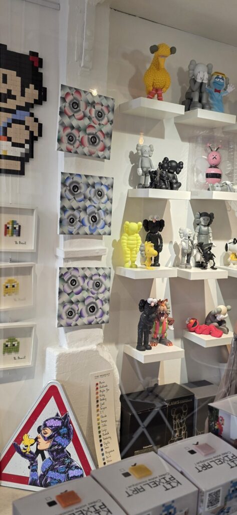 Gallery shelves crowded with vinyl figures including grey and black KAWS companions, a yellow Big Bird figure, pixel mosaic artworks and a Catwoman painting on a triangular road sign