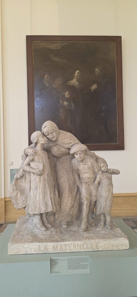 Large plaster sculpture group titled La Maternelle showing a woman in a shawl with four children pressed against her, standing in front of a big misty brown painting of figures