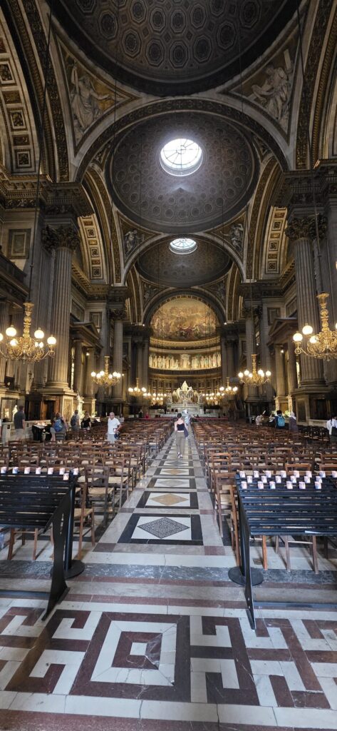 Interior of La Madeleine church in Paris looking down the aisle between rows of wooden chairs to the altar, with coffered domes overhead and a shaft of daylight coming through a round opening