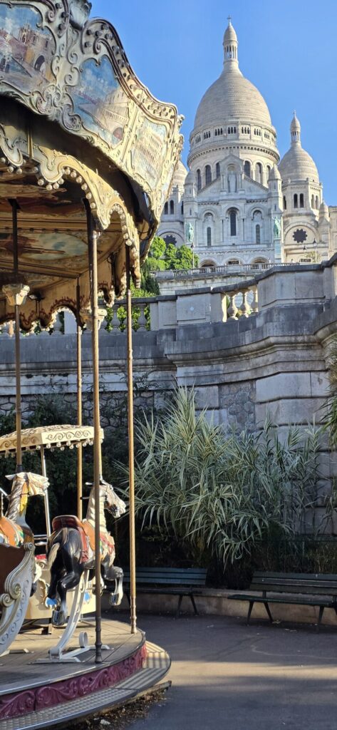 Old double decker carousel standing still and empty in morning shadow, painted panels and carved horses in the foreground, the white domes of the Sacre-Coeur rising over the stone balustrade behind it, empty green benches to the right