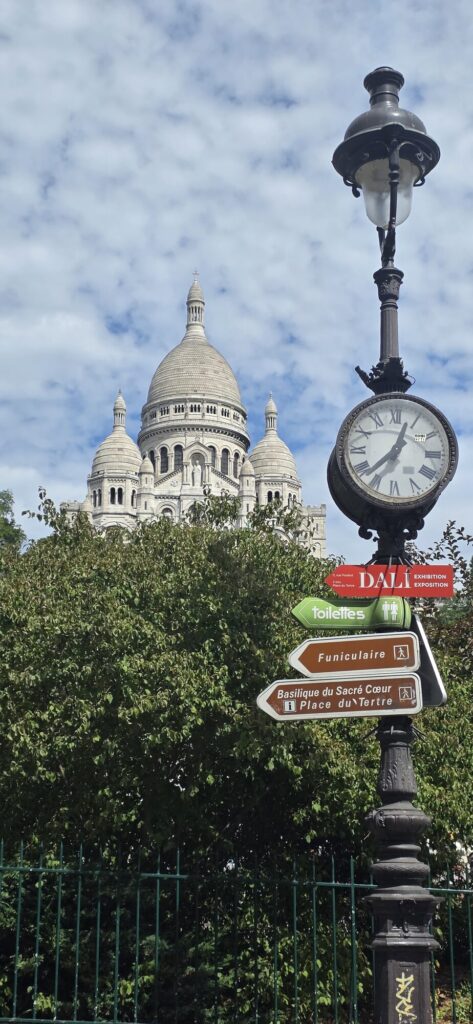 White domes of the Sacre-Coeur rising behind a dense green tree, with an ornate cast iron lamp post in front carrying a roman numeral clock and direction signs to the Dali exhibition, the funicular and the Place du Tertre
