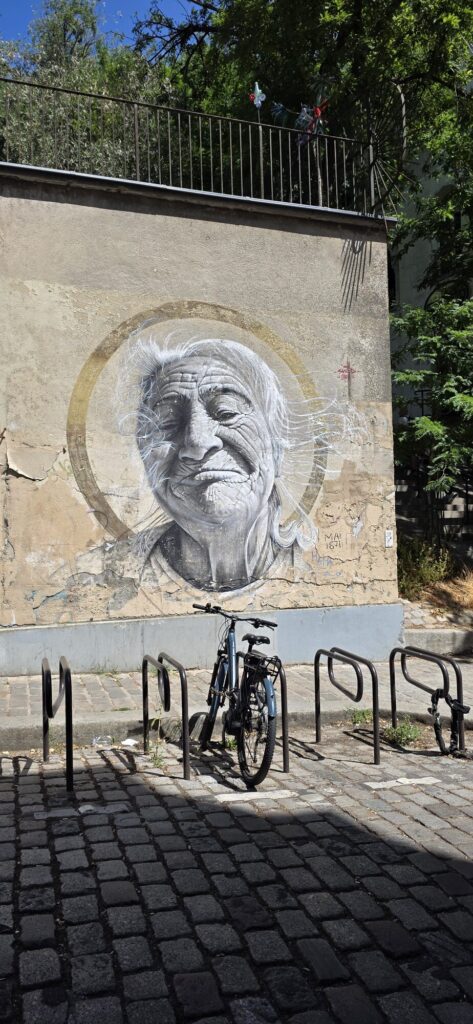 Large monochrome painted portrait of an elderly person with wild white hair inside a gold ring like a halo, filling a rendered wall above a row of empty bike racks on cobbles