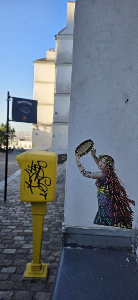 Empty cobbled Montmartre street in early sun with a bright yellow post box covered in black tags, and pasted on the white corner wall beside it a woman in a rainbow striped dress holding a tambourine above her head