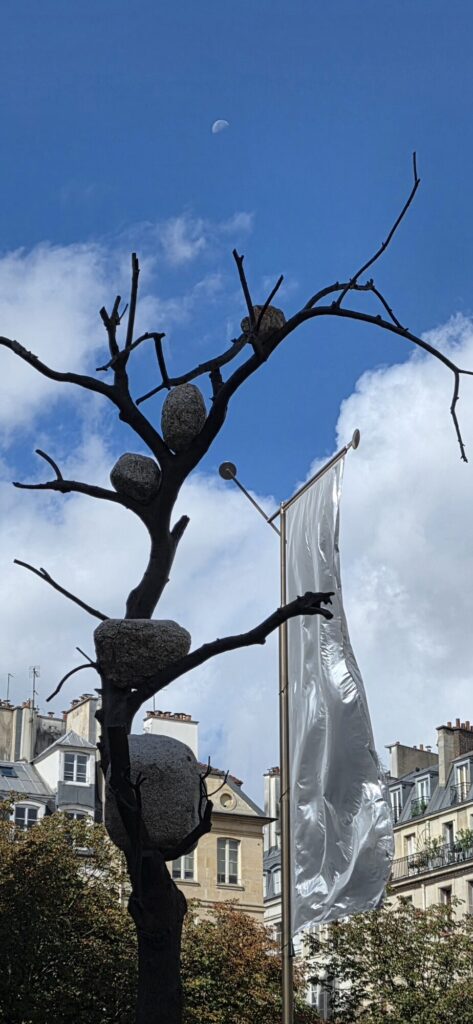 Bronze sculpture of a bare tree against a blue sky with white cloud, several large river stones lodged in the forks of its branches, a daytime half moon visible top left
