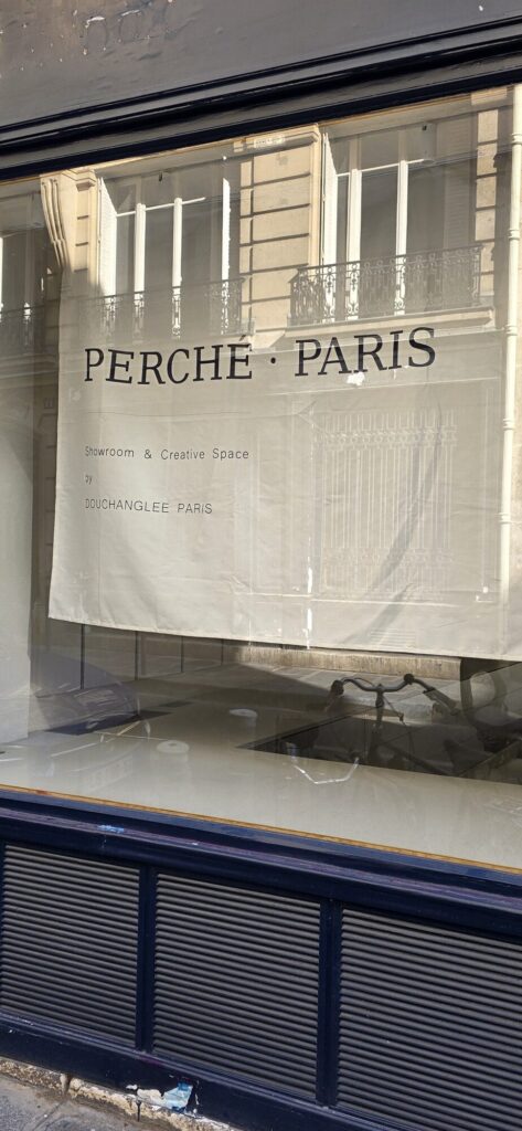 Paris shopfront with a large sheet of pale paper hung inside the window printed with the words Perche Paris, a dark blue shutter pulled down below it