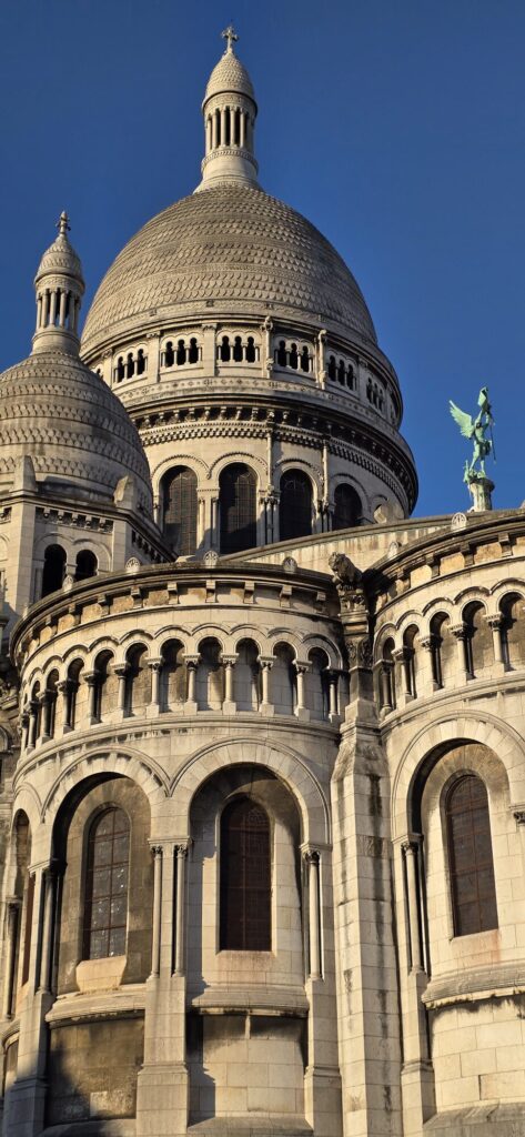 Close view of the white domes and apse of the Sacre-Coeur in low morning sun against a deep blue sky, with a green bronze angel on the roofline to the right and a small gargoyle below it
