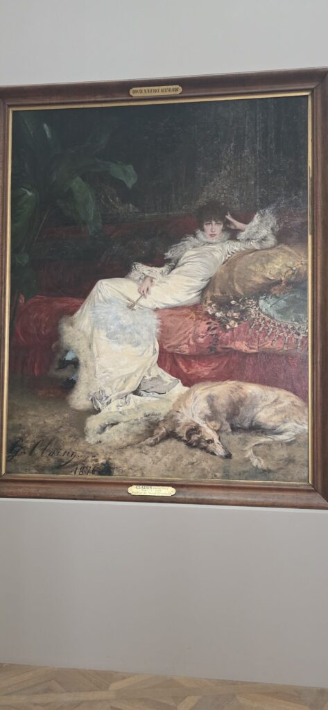 Georges Clairin's 1876 portrait of Sarah Bernhardt reclining in white satin on a red divan, with a large pale wolfhound stretched out asleep on the floor in front of her