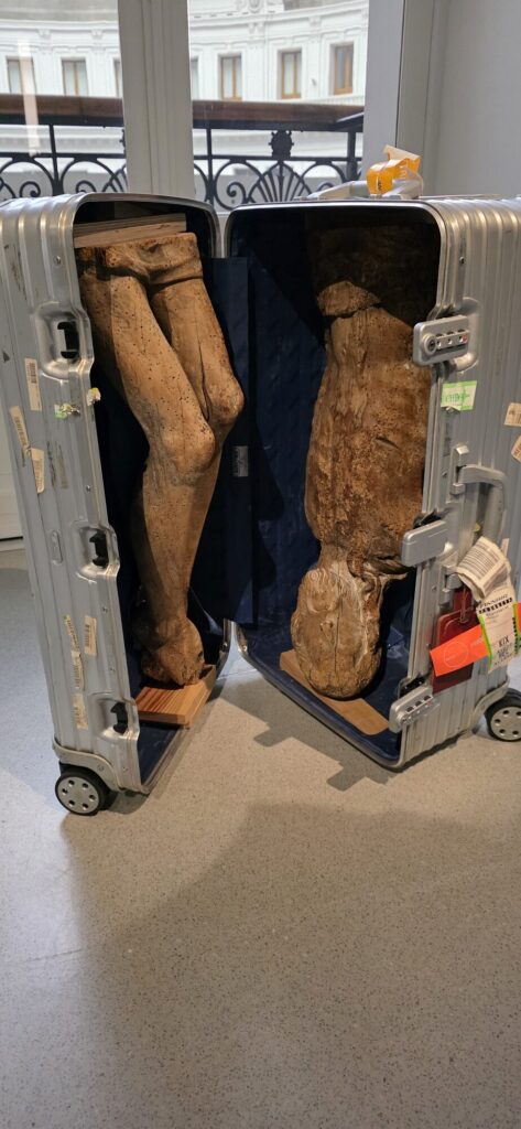 Battered aluminium flight case standing open on a gallery floor with a carved wooden figure packed inside it in two halves, legs in one side and torso in the other, old airline tags still tied to the handle