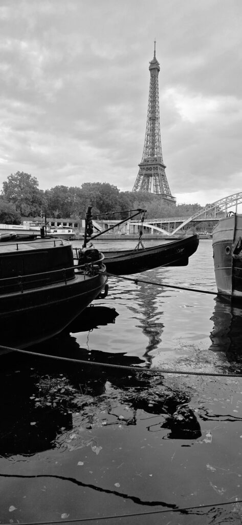 Black and white view along the Seine with the blunt bows of moored barges in the foreground, a mooring rope crossing the water, a footbridge behind and the Eiffel Tower on the skyline