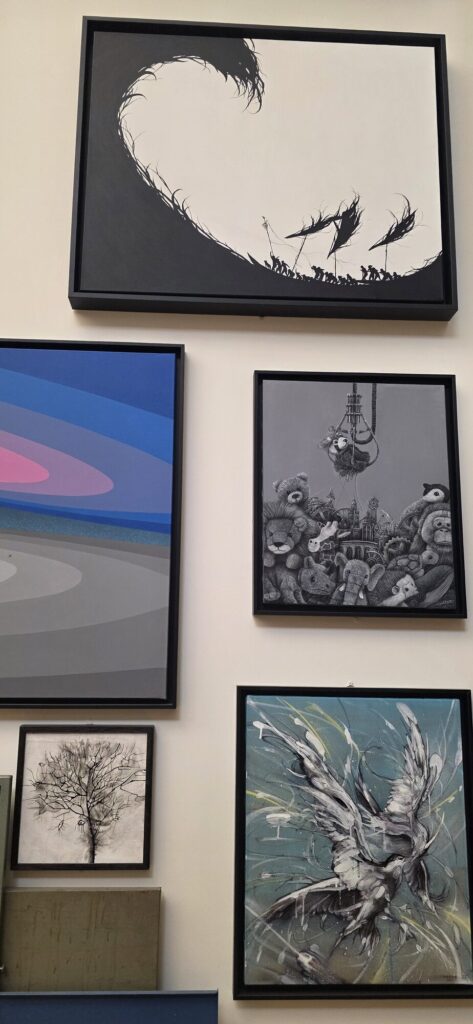 Gallery wall hung salon style with framed street art works including a large black and white brush painting of a curling wave with tiny figures below it, a fine pencil drawing of a bare tree, and a painted bird in white strokes
