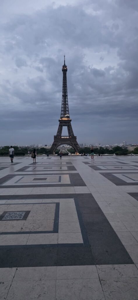 The wide stone esplanade at Trocadero at dawn under heavy grey cloud, its dark and pale paving laid in large interlocking rectangular patterns, with the Eiffel Tower standing beyond
