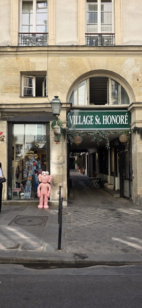 Stone passage arch with a green Village Saint Honore sign, a hanging lantern and planters, and a two metre pink furry KAWS companion figure standing at a gallery window on the right